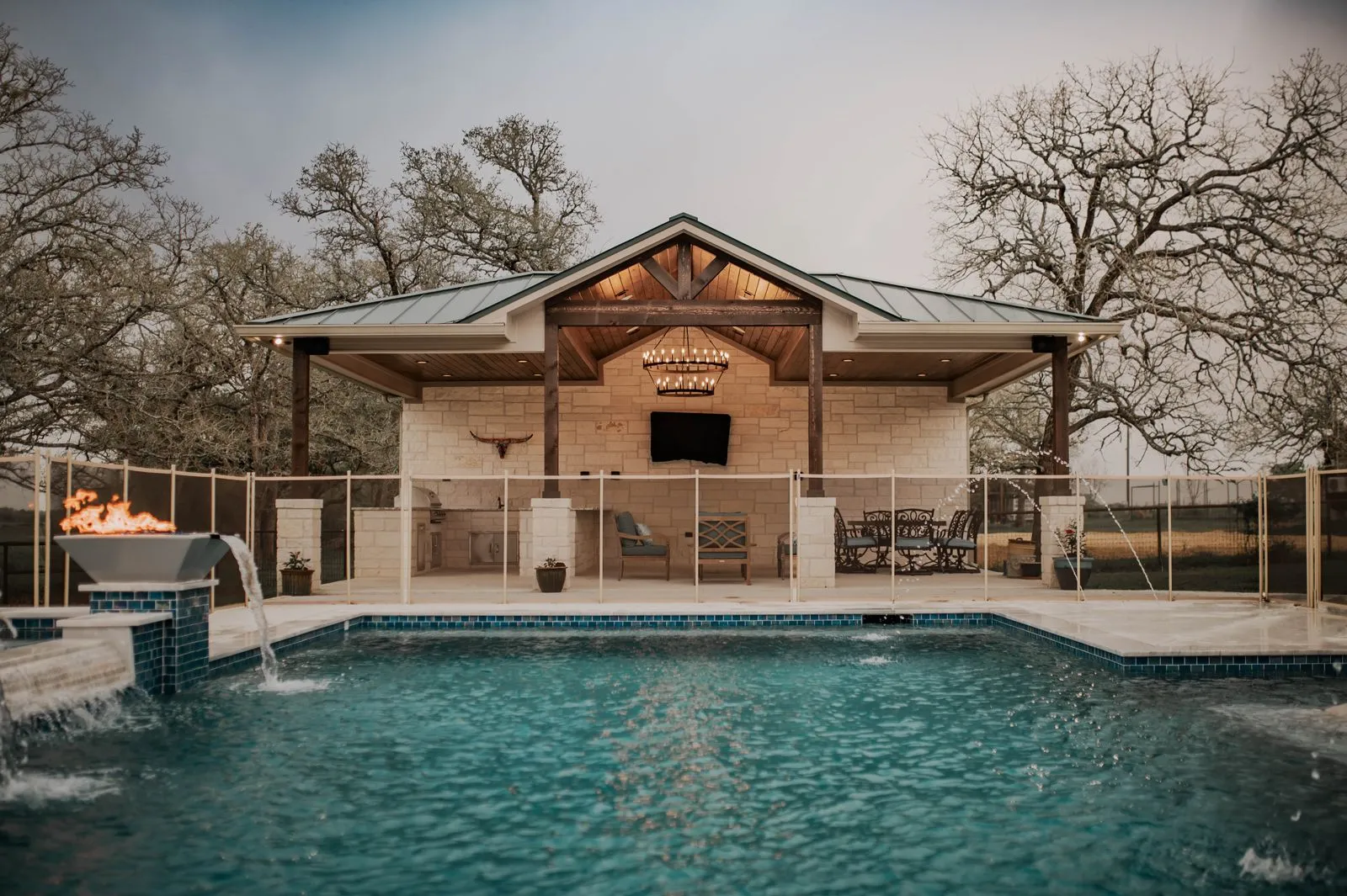 A luxurious outdoor pool area features a modern pavilion with a stone facade, elegant lighting, and a shimmering pool, surrounded by trees
