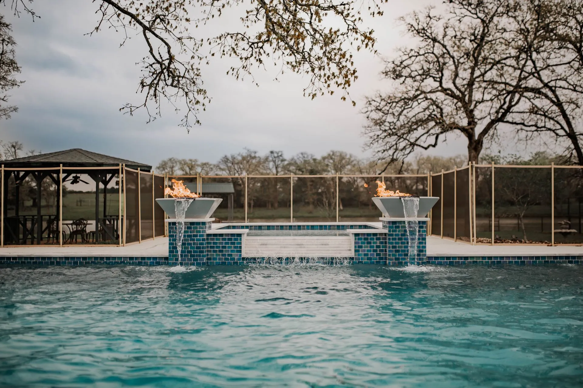 A luxurious pool features two flame-lit fountains, surrounded by a safety fence and a shaded seating area in a serene outdoor setting A luxurious pool features two flame-lit fountains, surrounded by a safety fence and a shaded seating area in a serene outdoor setting