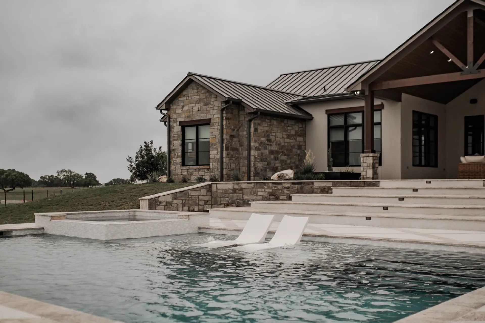 A modern stone house with a pool featuring two white loungers, set against a cloudy sky and surrounded by lush greenery A modern stone house with a pool featuring two white loungers, set against a cloudy sky and surrounded by lush greenery