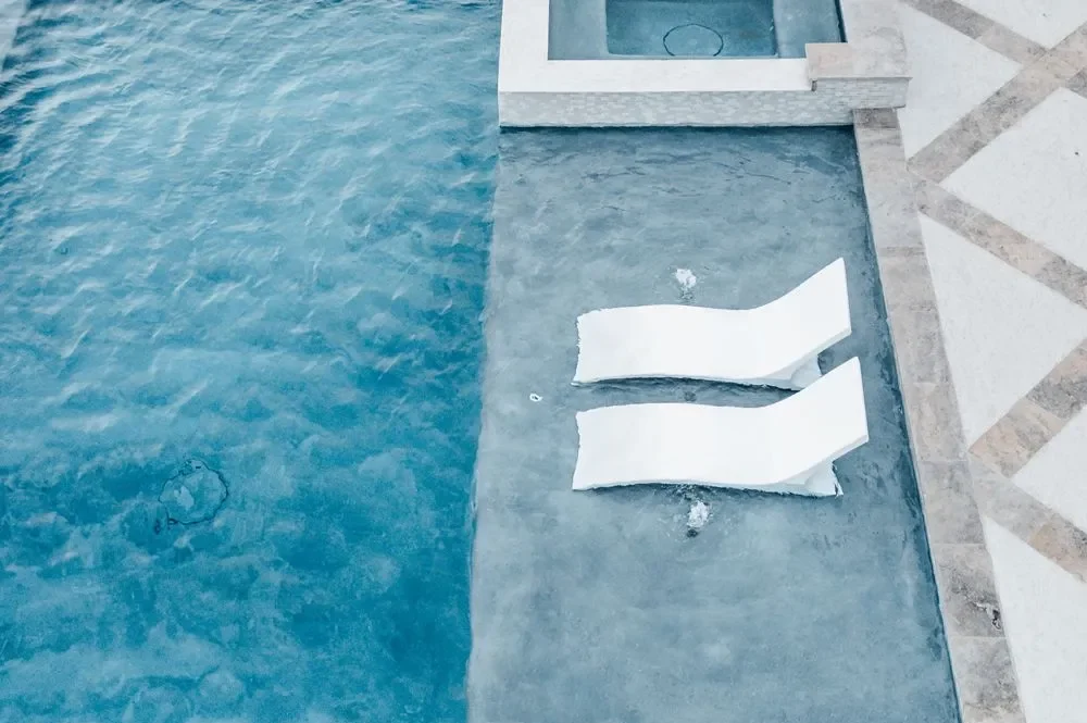 Aerial view of two white lounge chairs partially submerged in a clear blue swimming pool surrounded by a textured stone deck Aerial view of two white lounge chairs partially submerged in a clear blue swimming pool surrounded by a textured stone deck
