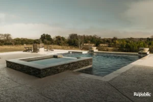A serene outdoor pool area featuring a hot tub, stone accents, and wooden chairs, surrounded by lush landscaping under a soft sky