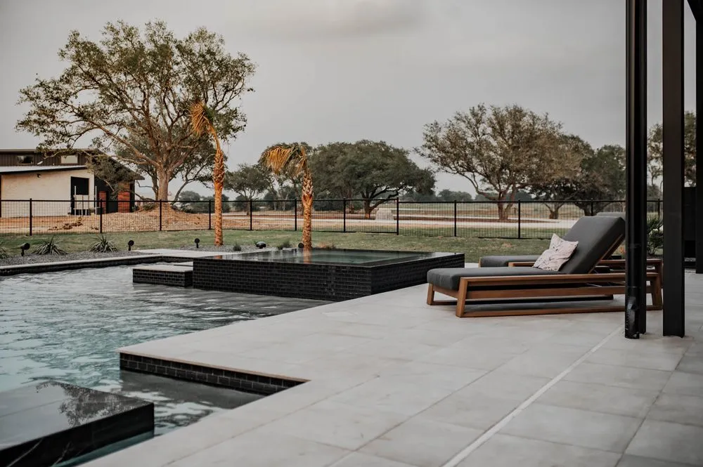 Modern patio with a pool and lounge chair, framed by palm trees and open grassy landscape under an overcast sky Modern patio with a pool and lounge chair, framed by palm trees and open grassy landscape under an overcast sky